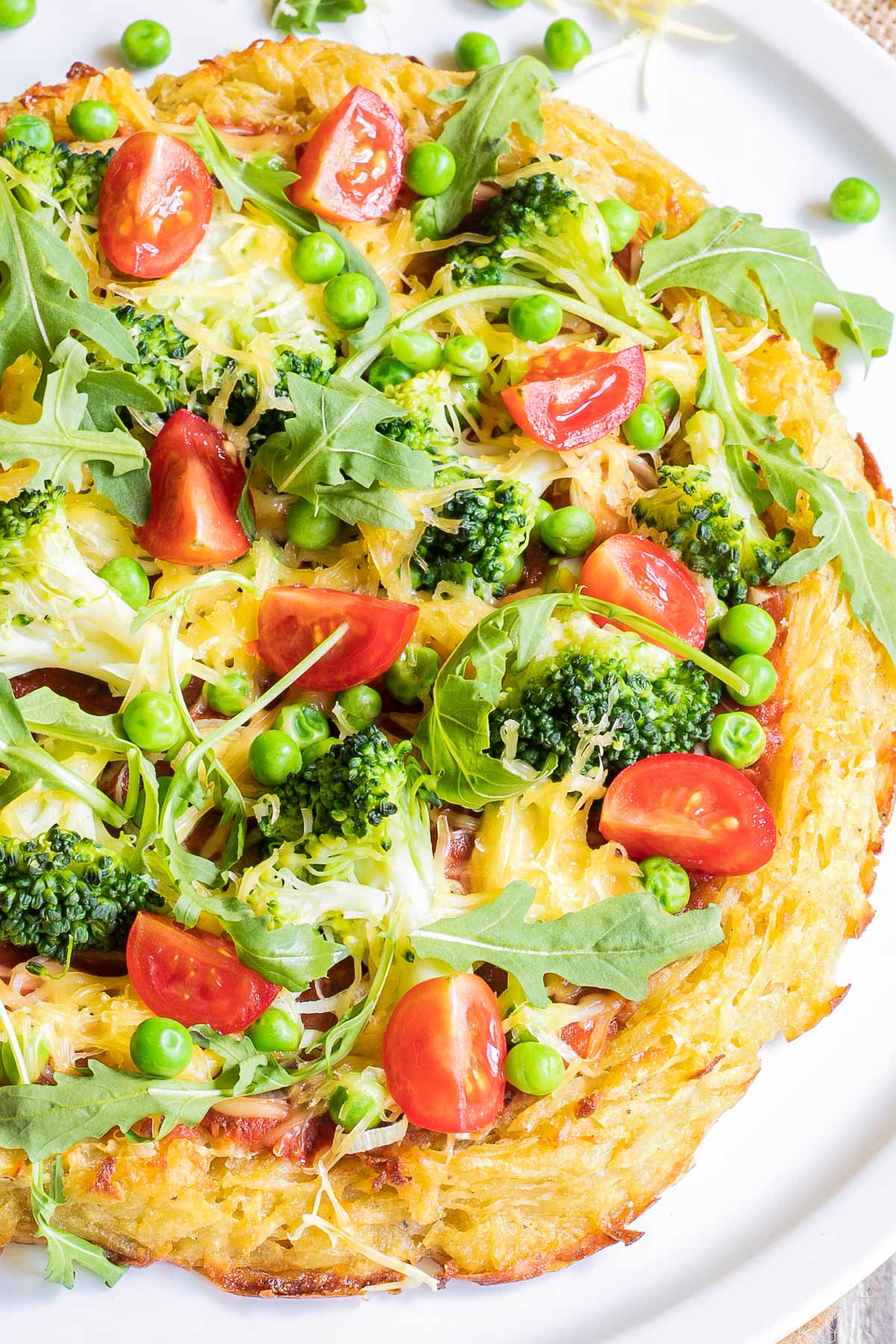 Hash brown crust pizza with topped with tomato sauce, broccoli, peas cherry tomatoes, arugula, and shredded melted cheese. Served on a white plate.