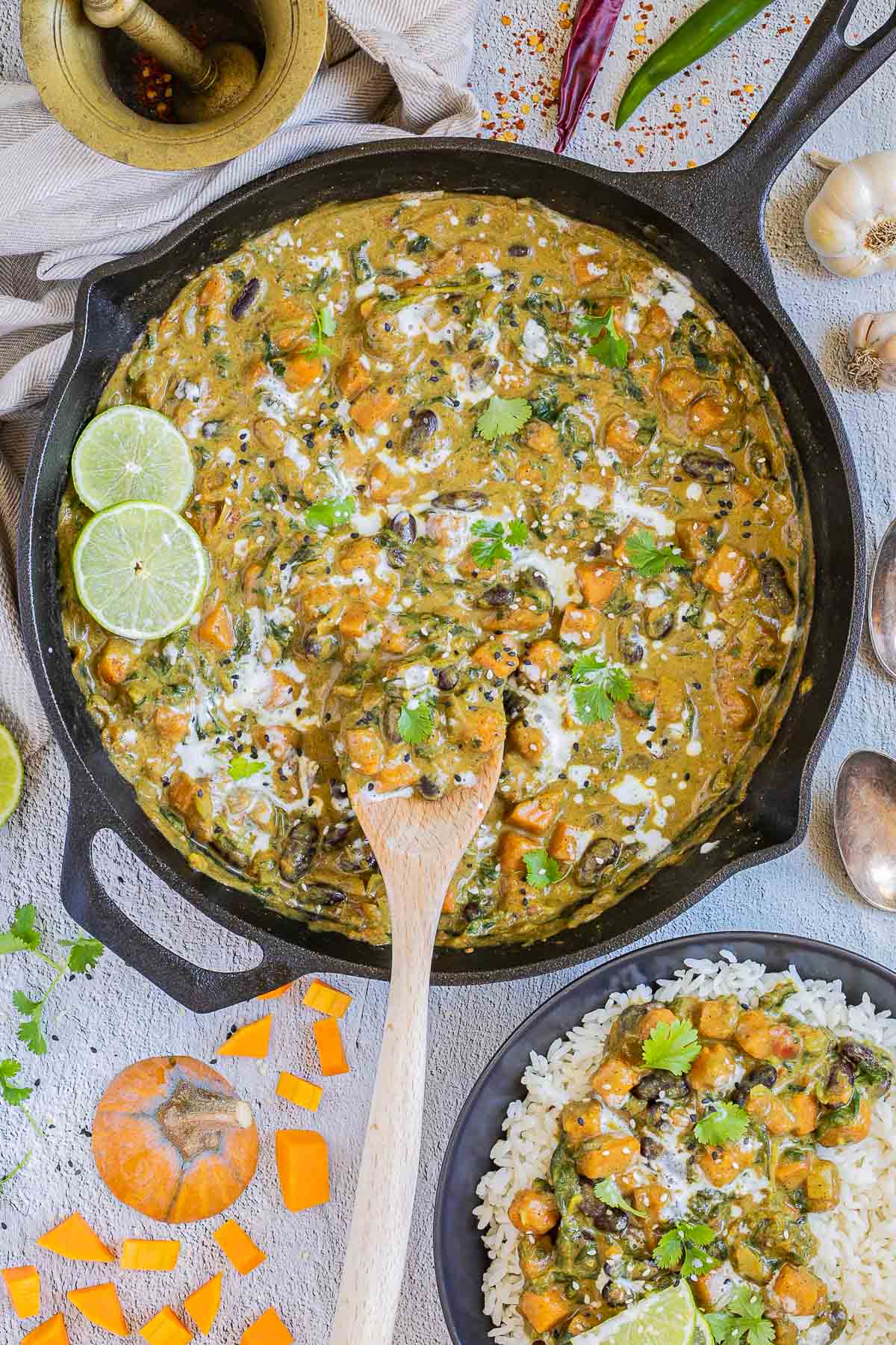 Cast iron skillet with a green-brown curry sauce, orange butternut squash cubes, red kidney beans, and spinach pieces. It is drizzled with white sauce and topped with fresh green herbs. A small bowl with rice and curry is next to it.