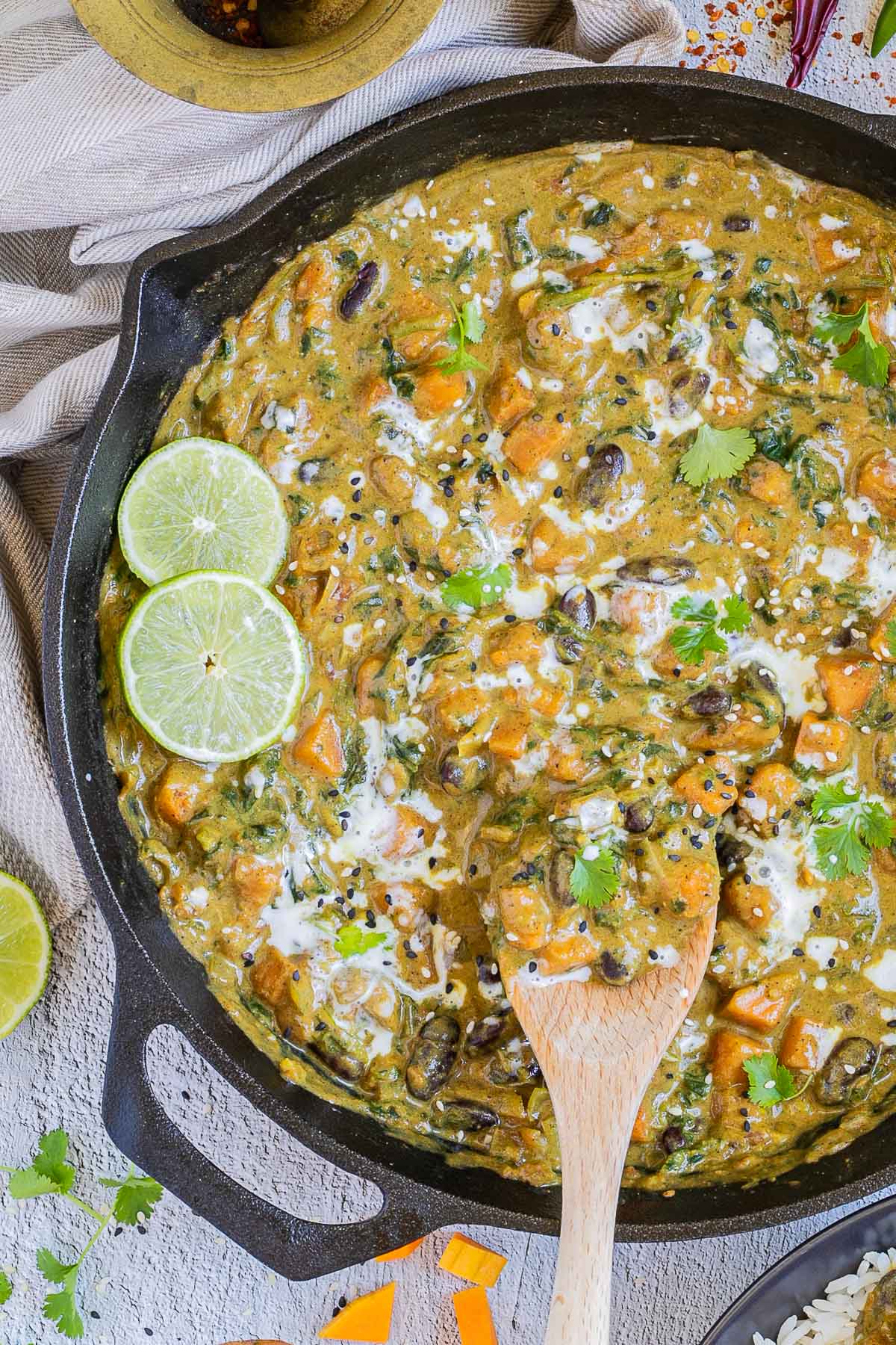 Cast iron skillet with a green-brown curry sauce, orange butternut squash cubes, red kidney beans, and spinach pieces. It is drizzled with white sauce and topped with fresh green herbs. A wooden spatula is placed inside.