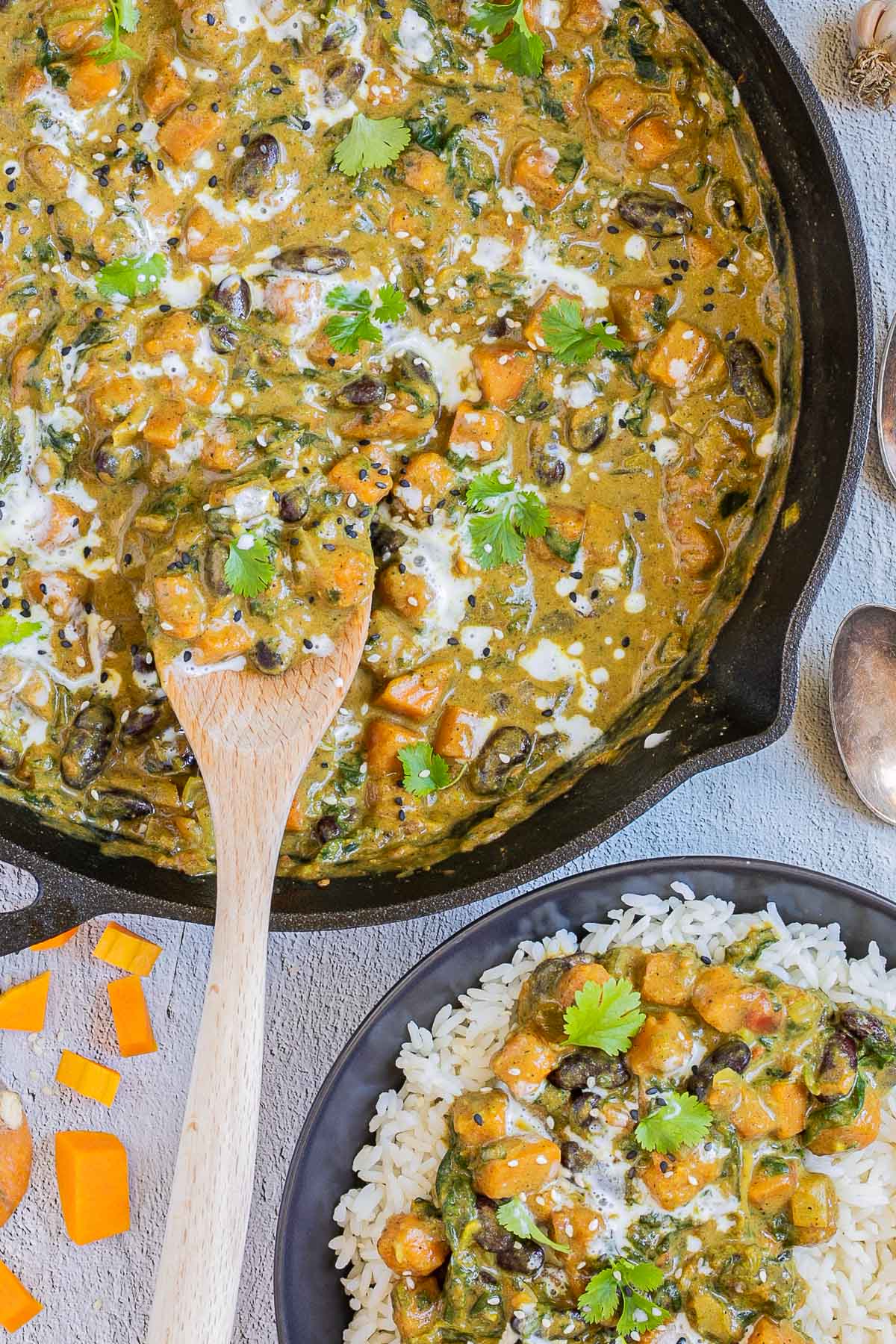 Cast iron skillet with a green-brown curry sauce, orange butternut squash cubes, red kidney beans, and spinach pieces. It is drizzled with white sauce and topped with fresh green herbs. A small bowl with rice and curry is next to it.