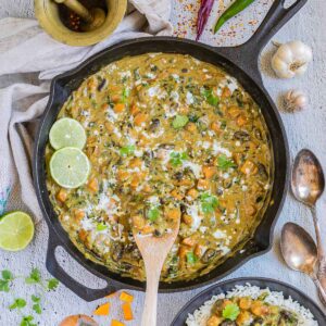 Cast iron skillet with a green-brown curry sauce, orange butternut squash cubes, red kidney beans, and spinach pieces. It is drizzled with white sauce and topped with fresh green herbs. A small bowl with rice and curry is next to it.