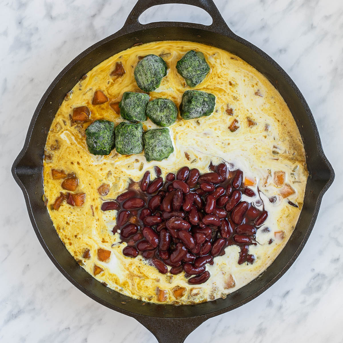 Light orange sauce with orange squash cubes, red kidney beans and frozen spinach leaves in a cast iron skillet