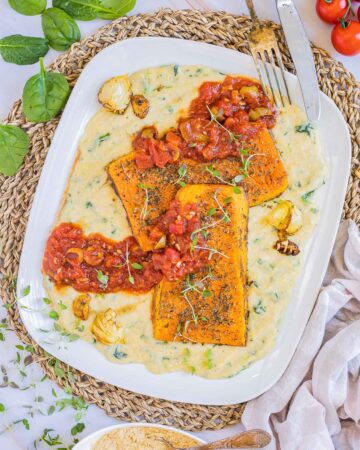 Large white plate with creamy polenta and large butternut squash slices drizzled with red tomato sauce and topped with fresh herbs.