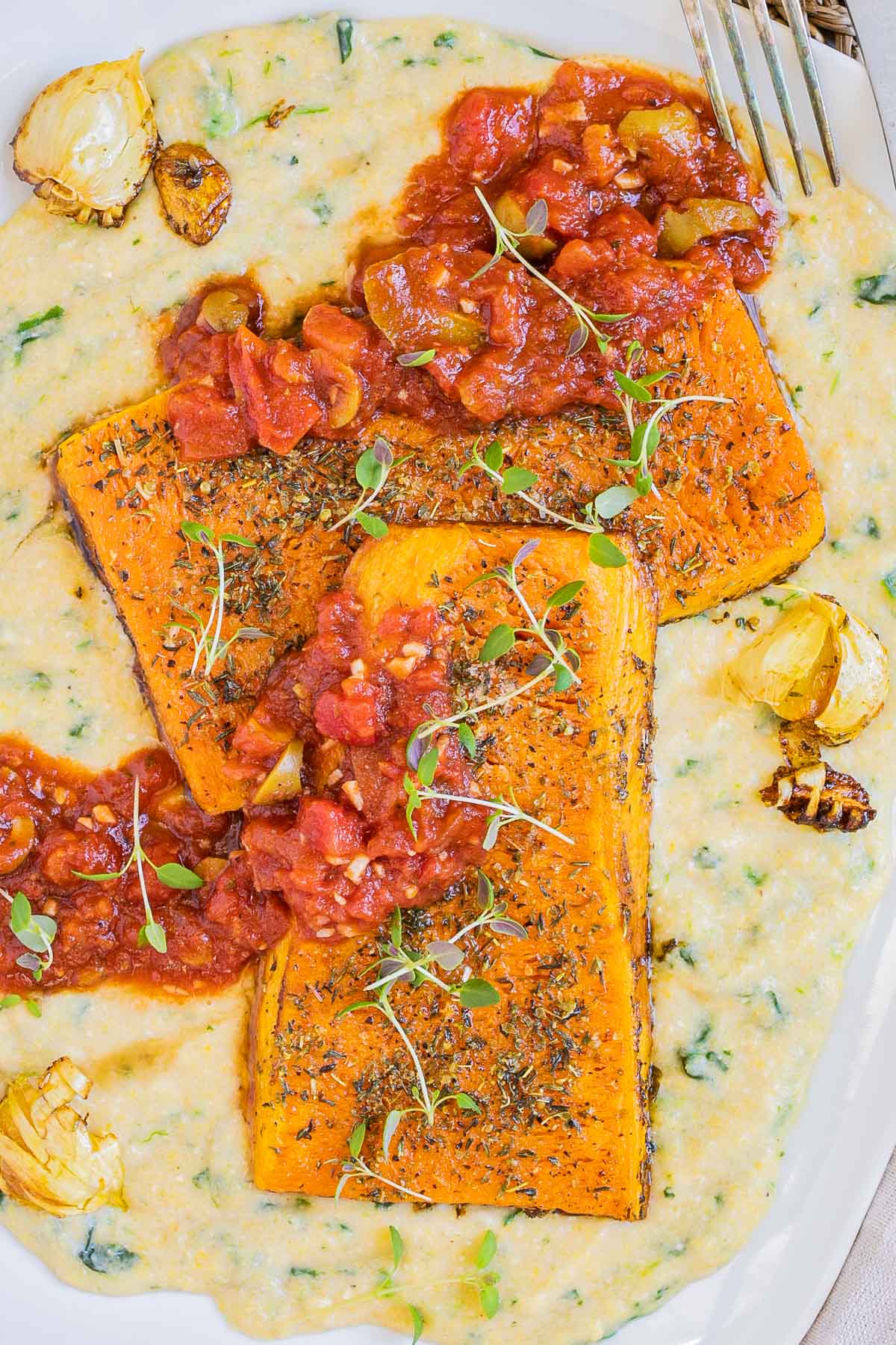 Large white plate with creamy polenta and large butternut squash slices drizzled with red tomato sauce and topped with fresh herbs.