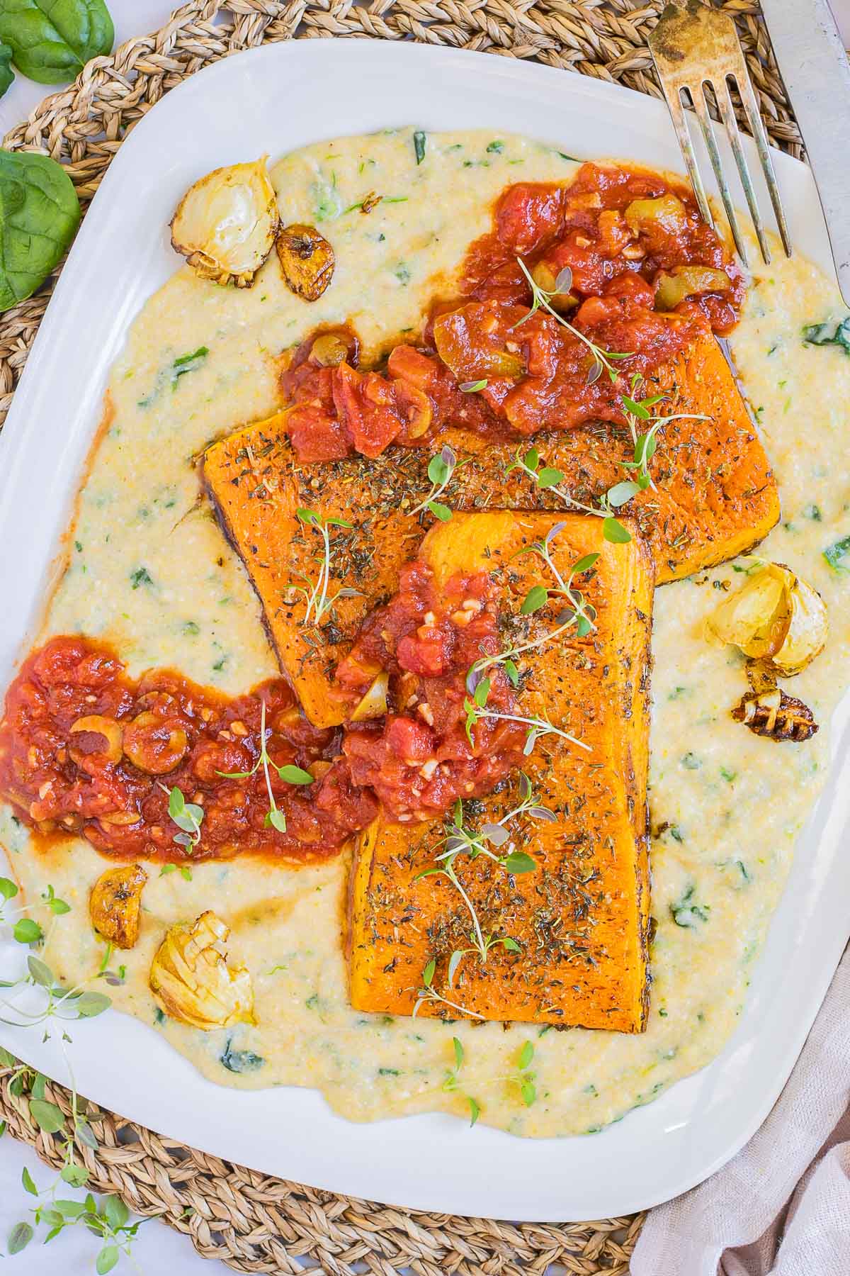 Large white plate with creamy polenta and large butternut squash slices drizzled with red tomato sauce and topped with fresh herbs.