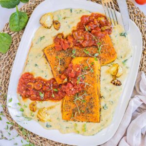 Large white plate with creamy polenta and large butternut squash slices drizzled with red tomato sauce and topped with fresh herbs.