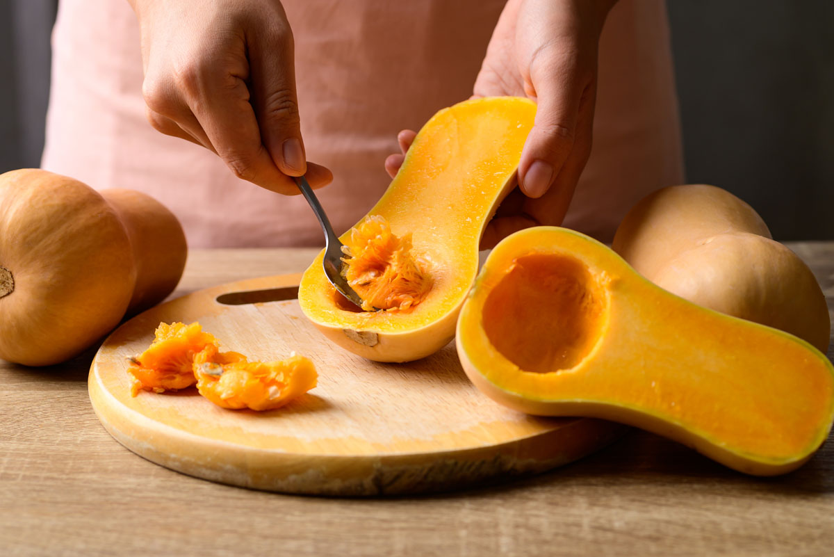 A hand is holding half butternut squash while th other hand is scooping the seeds out.  