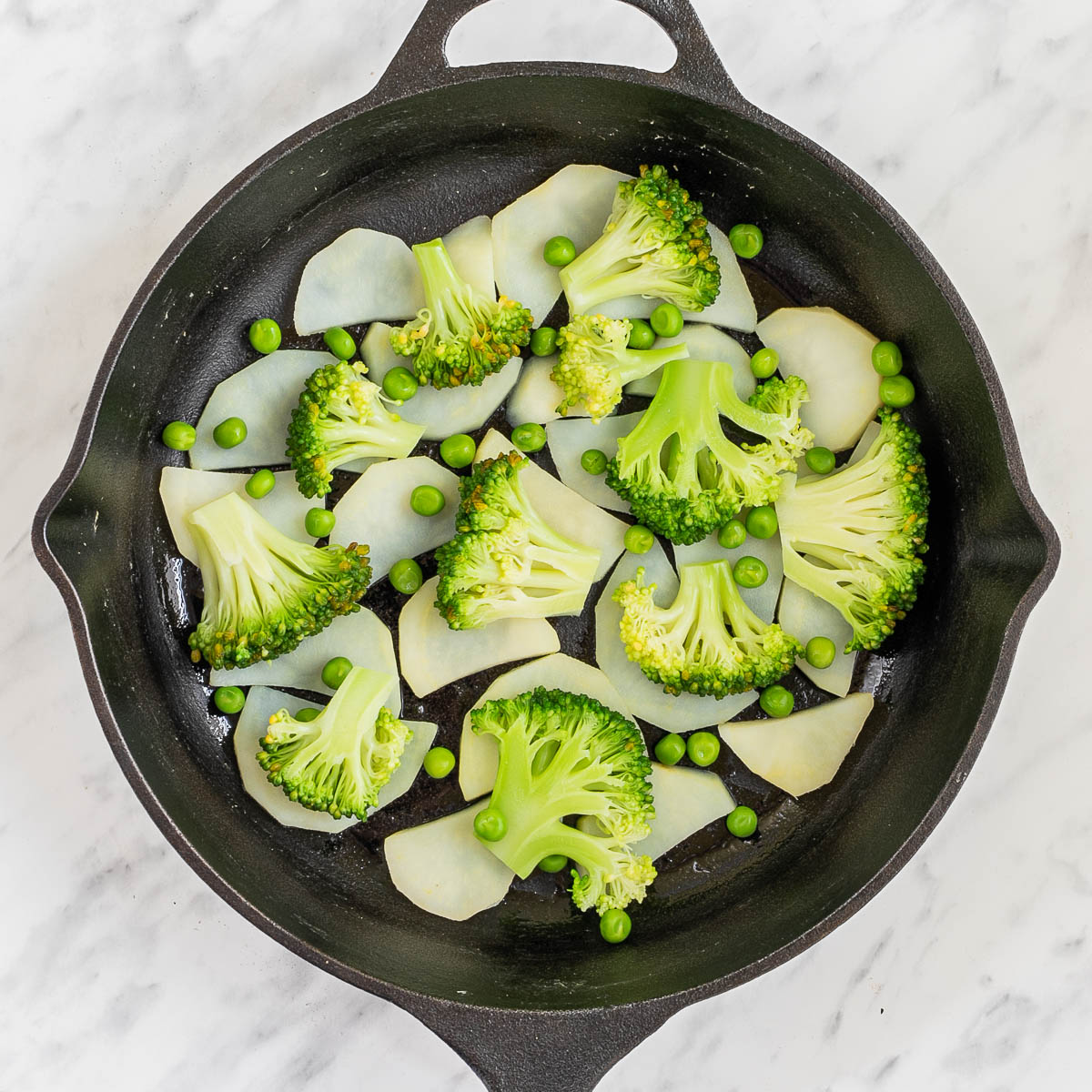Black cast iron skillet with potato slices, broccoli, and green peas.
