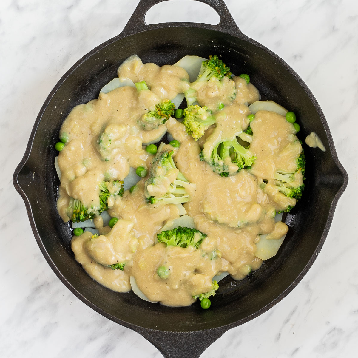 Black cast iron skillet with potato slices, broccoli, and green peas. topped with yellow thick sauce.