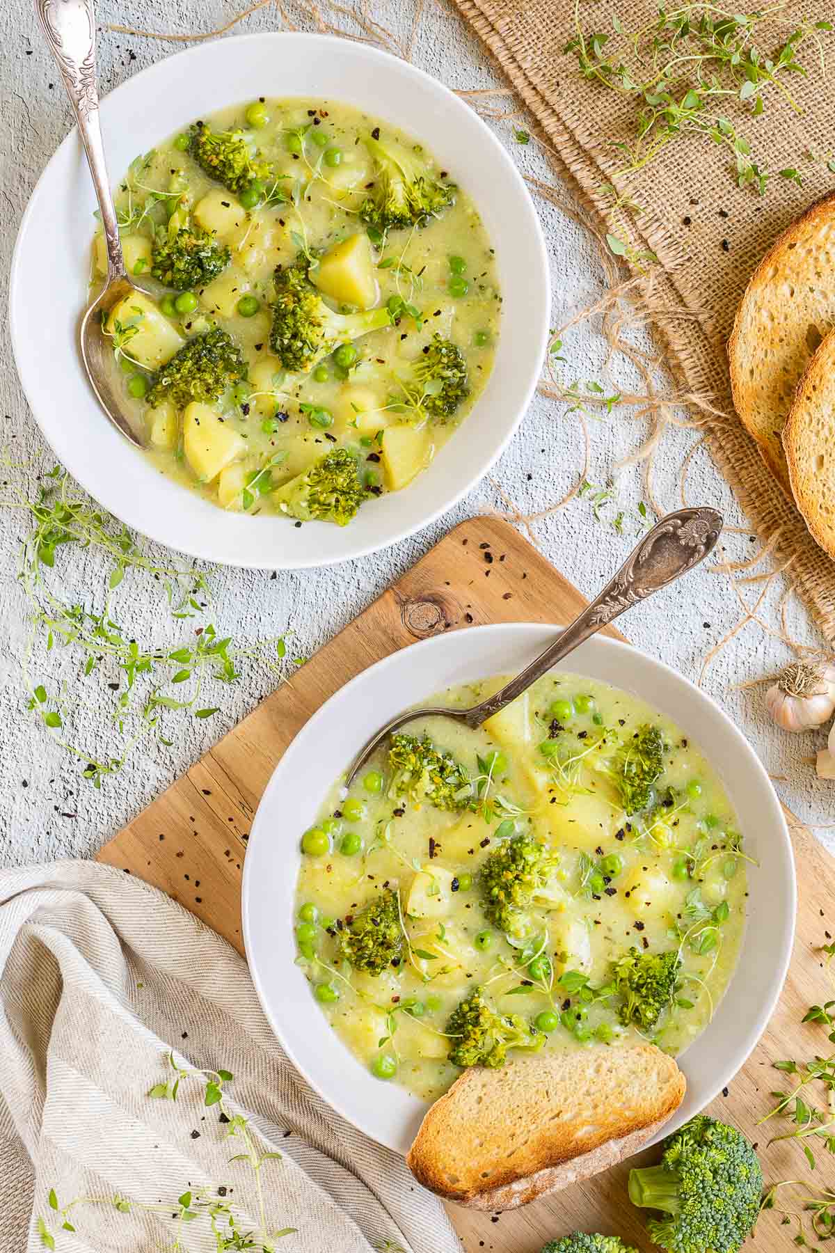 2 white plates of thick yellow stew with potatoes, broccoli, green peas and green herbs. A spoon is placed inside with a toasted slice of bread.