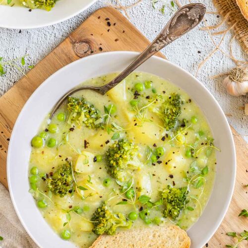 2 white plates of thick yellow stew with potatoes, broccoli, green peas and green herbs. A spoon is placed inside with a toasted slice of bread.