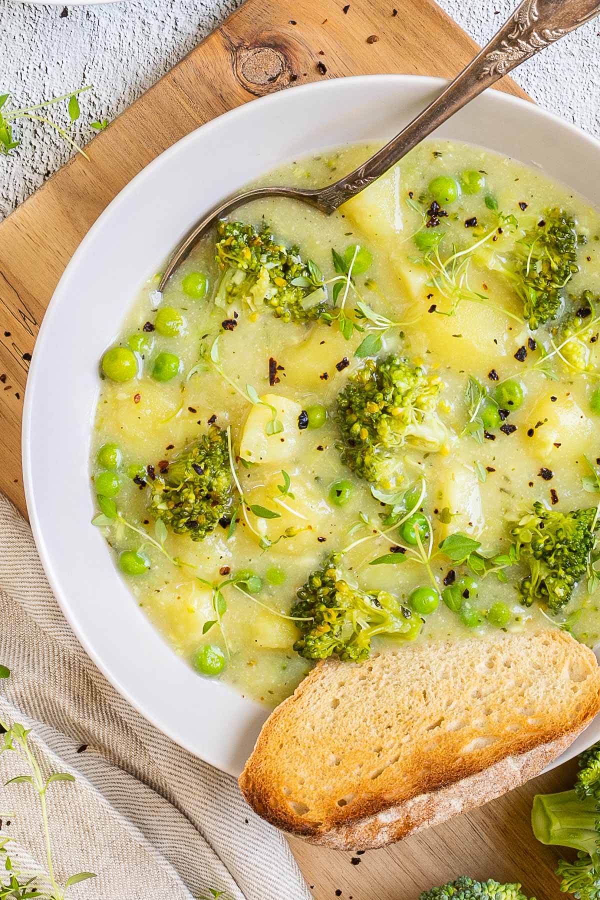 A white plate of thick yellow stew with potatoes, broccoli, green peas and green herbs. A spoon is placed inside with a toasted slice of bread.