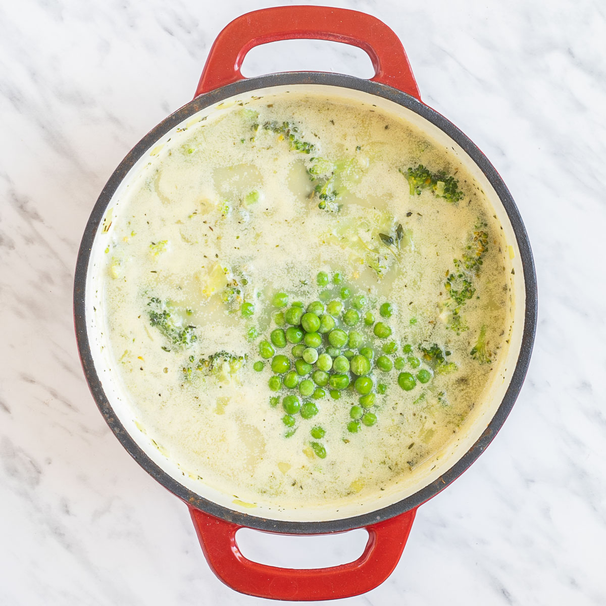 Red and white enameled Dutch oven with yellow stew of large potato chunks, broccoli, leek slices, green peas, and green herbs.