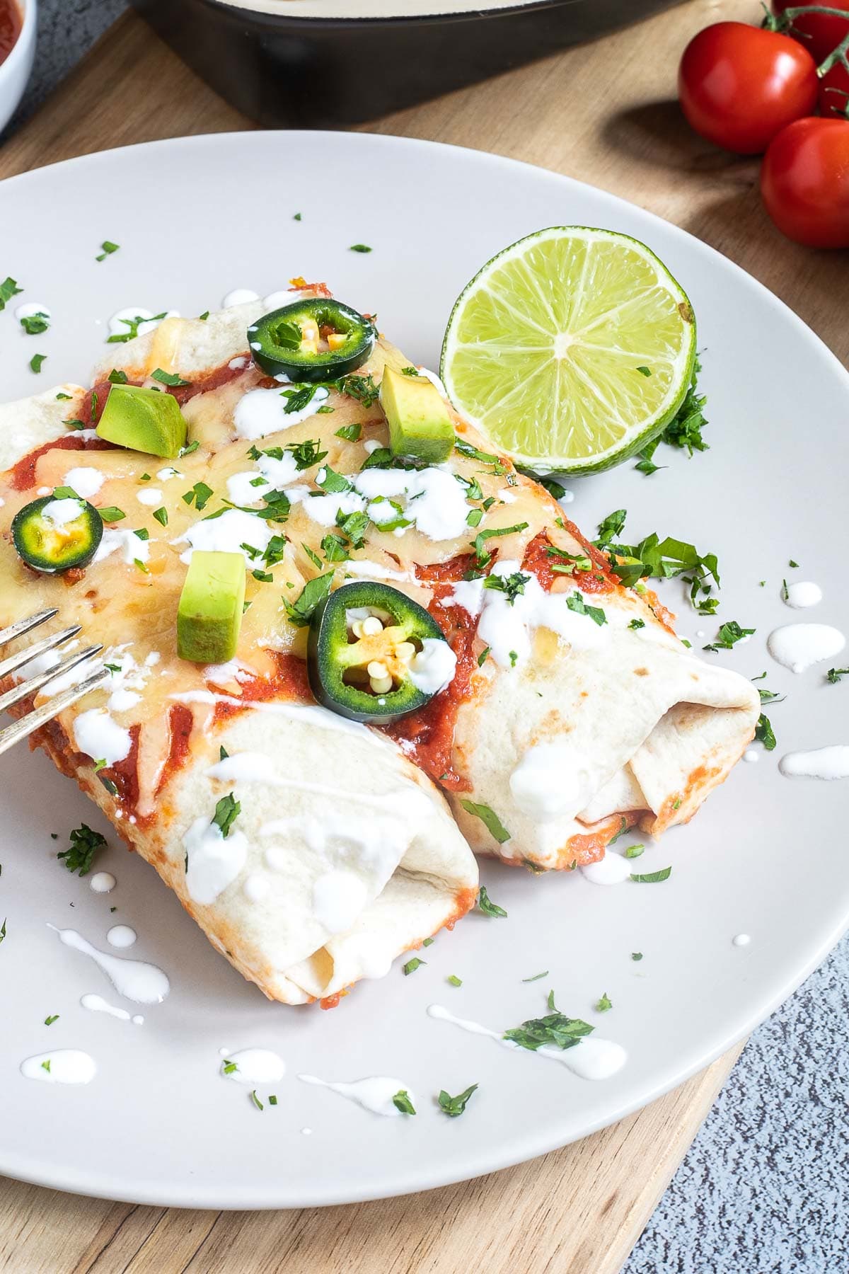 2 tortilla burrito on a white plate topped with red sauce, melted cheese, sour cream, green chili peppers and fresh chopped herbs. Remaining tortillas are in a pan next to it.