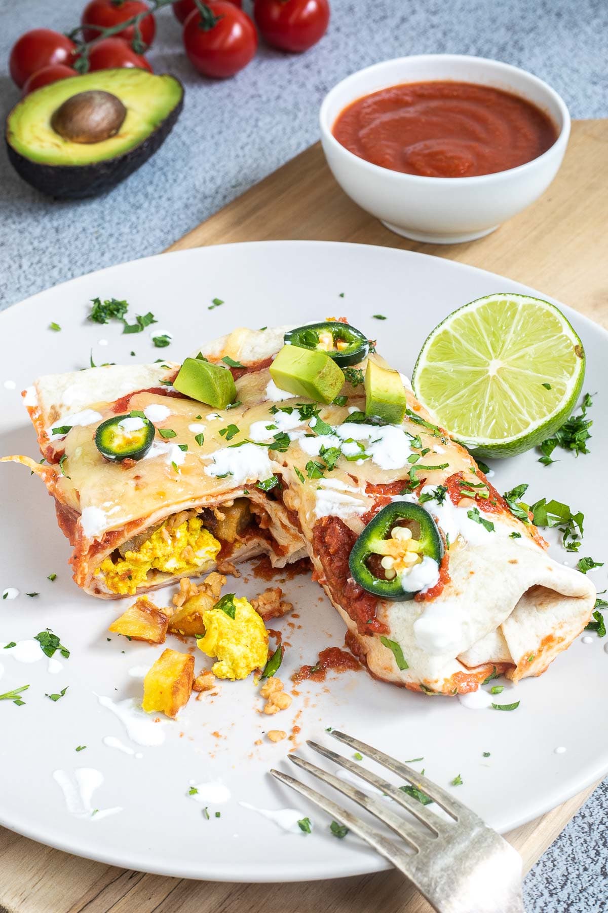 2 tortilla burrito on a white plate topped with red sauce, melted cheese, sour cream, green chili peppers and fresh chopped herbs. One is cut in half and a yellow filling is visible.