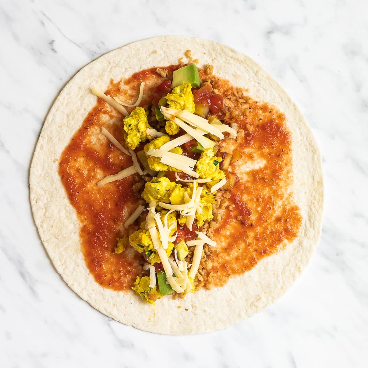 A tortilla on a marble surface covered partly with red sauce and topped with sausage crumbles, diced potatoes, avocado, and tofu scrambles.