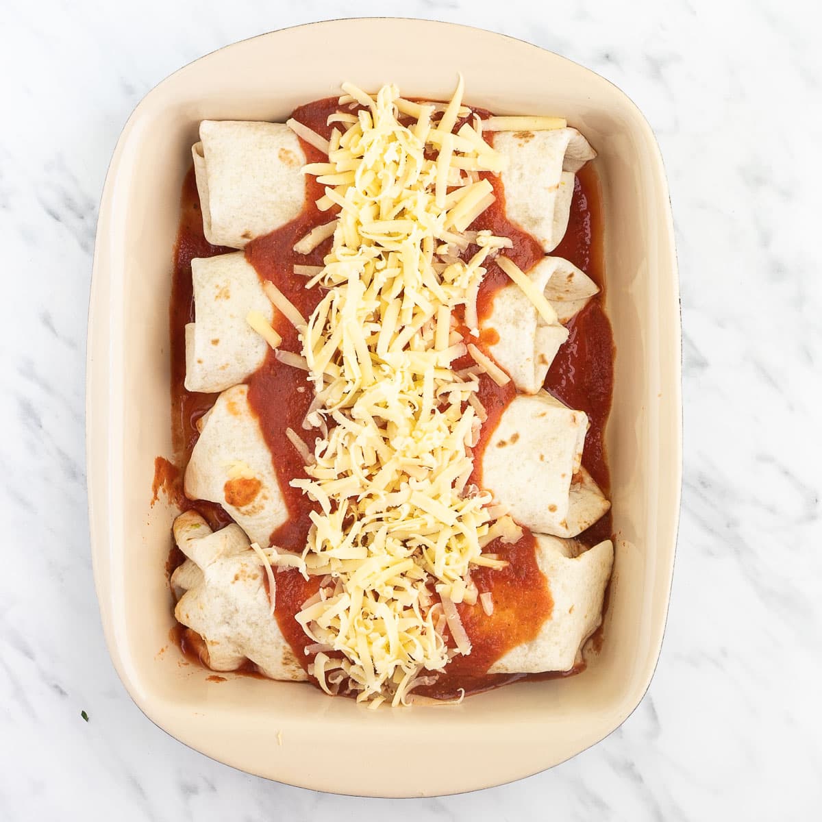 4 tortilla wraps in a red sauce topped with more red sauce and shredded cheese tucked closely together in a ceramic pan.