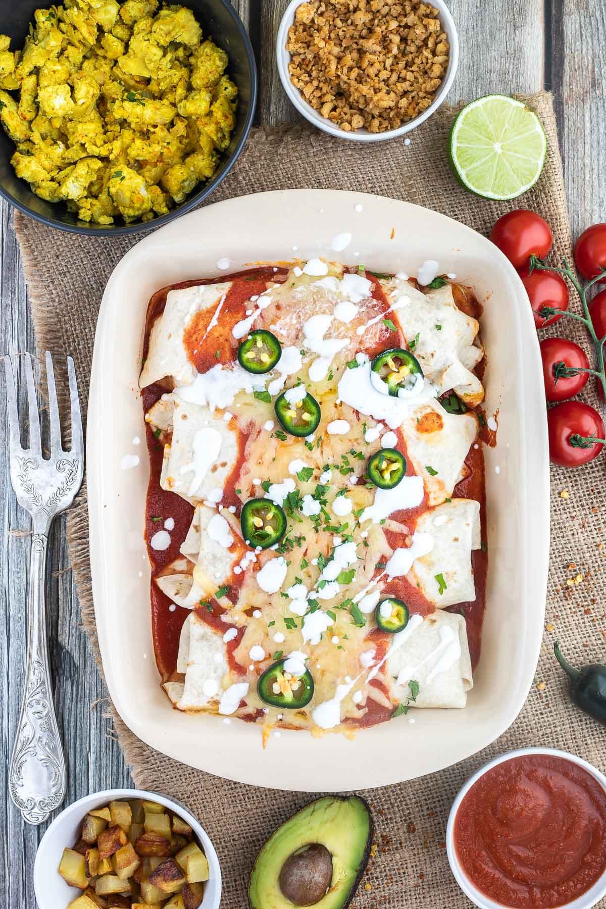 4 tortilla wraps topped with red sauce, melted cheese, sour cream, green chili peppers and fresh chopped herbs tucked closely together in a ceramic pan. Leftover filling ingredients are next to the pan in small bowls.