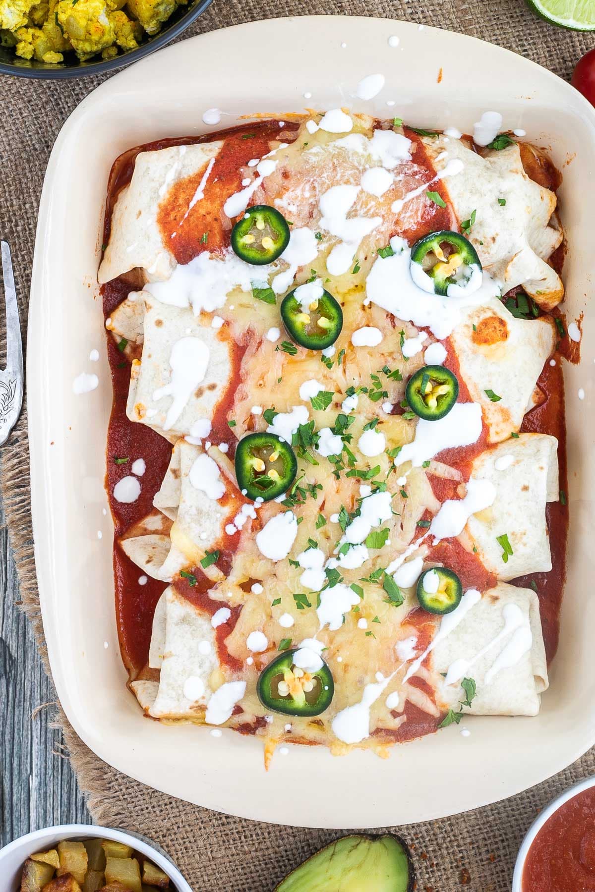 4 tortilla wraps topped with red sauce, melted cheese, sour cream, green chili peppers and fresh chopped herbs tucked closely together in a ceramic pan. Leftover filling ingredients are next to the pan in small bowls.