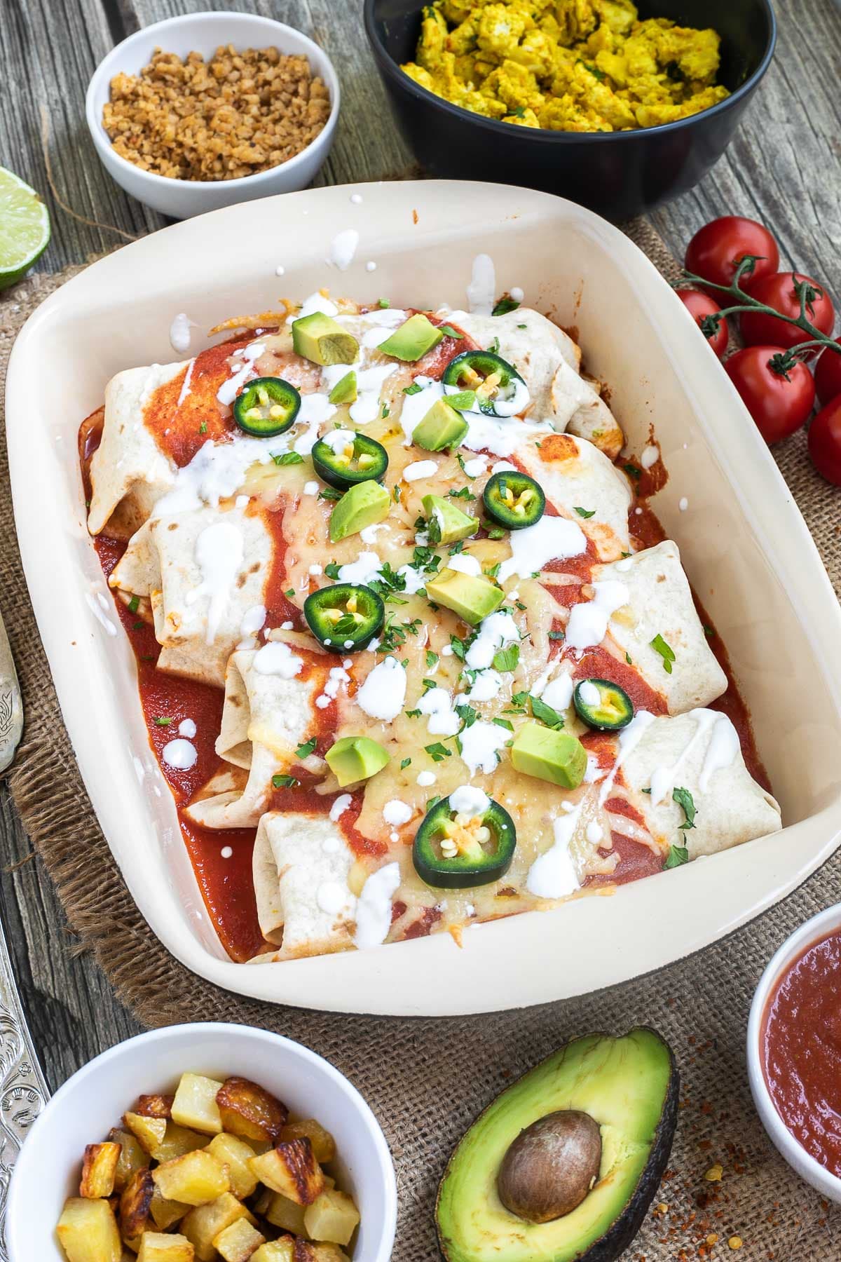 4 tortilla wraps topped with red sauce, melted cheese, sour cream, green chili peppers and fresh chopped herbs tucked closely together in a ceramic pan. Leftover filling ingredients are next to the pan in small bowls.