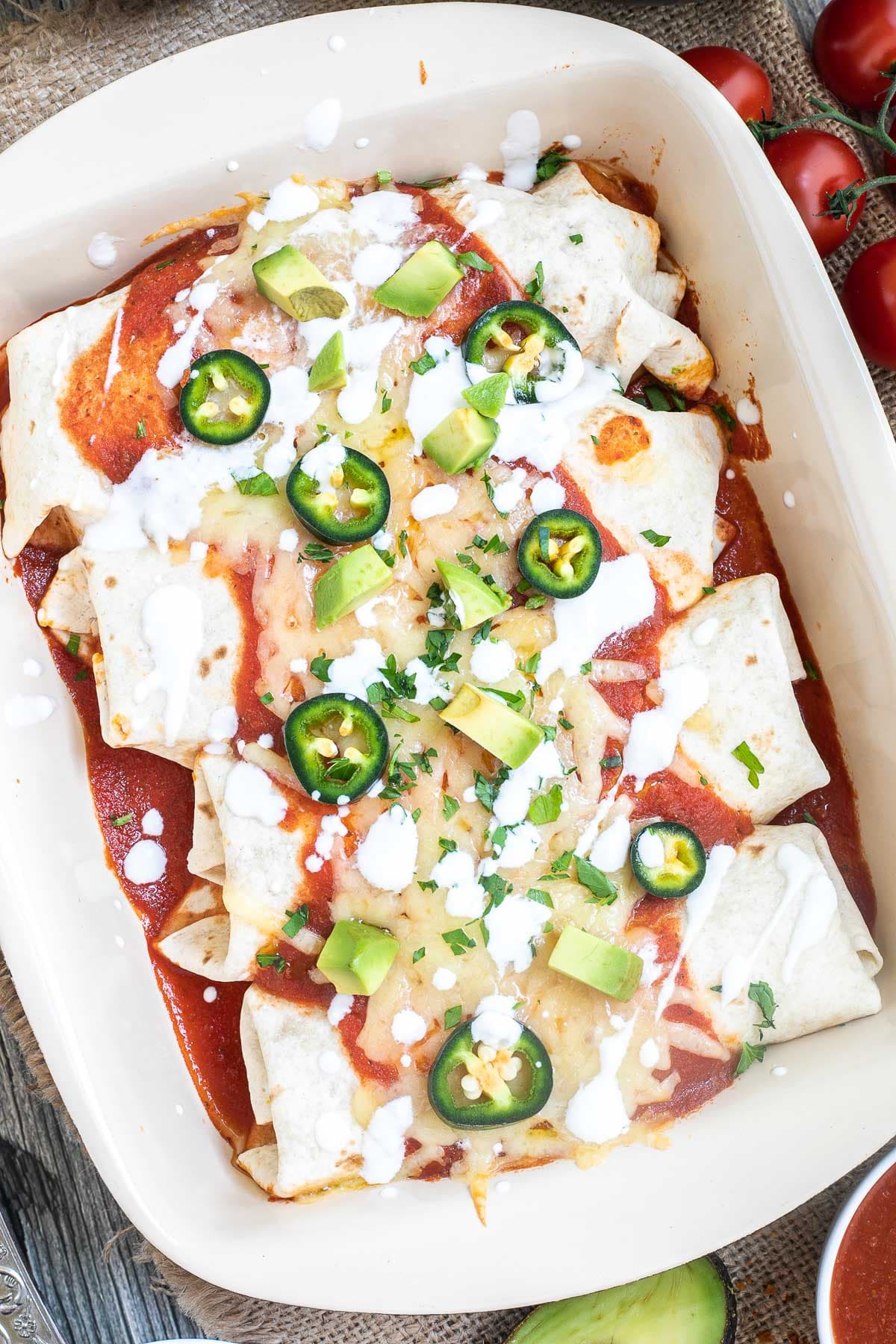 4 tortilla wraps topped with red sauce, melted cheese, sour cream, green chili peppers and fresh chopped herbs tucked closely together in a ceramic pan. Leftover filling ingredients are next to the pan in small bowls.