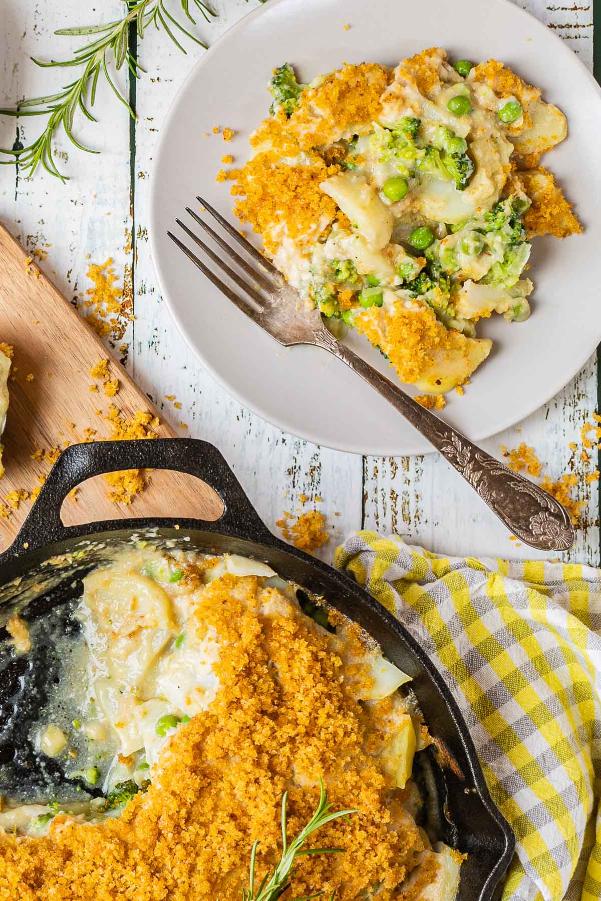 A black cast iron skillet with potato slices in a yellow thick sauce topped with yellow breadcrumbs. A part of it is missing and there is a gray plate with some food on it where you can see peas and broccoli as well.