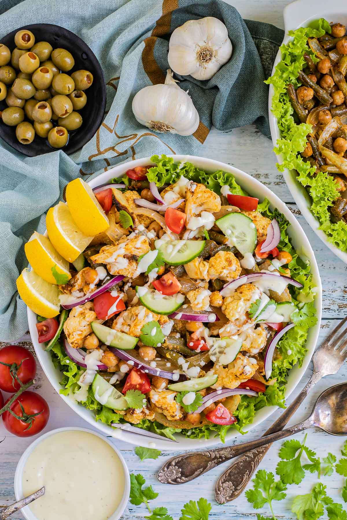 A large white bowl with green salad leaves topped with cauliflower florets, green beans, chickpeas, fresh tomatoes drizzled with a white sauce. Olives and more roasted veggies are next to it on small plates. 