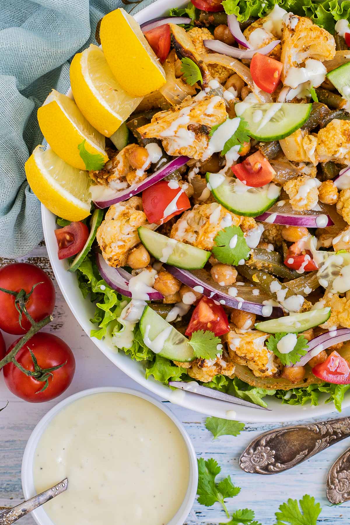 A large white bowl with green salad leaves topped with cauliflower florets, green beans, chickpeas, fresh tomatoes drizzled with a white sauce. Olives and more roasted veggies are next to it on small plates.