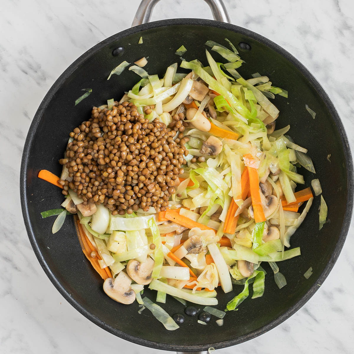 A wok with a mix of sliced mushrooms, shredded cabbage and sliced carrots. A small heap of brown lentils are added.