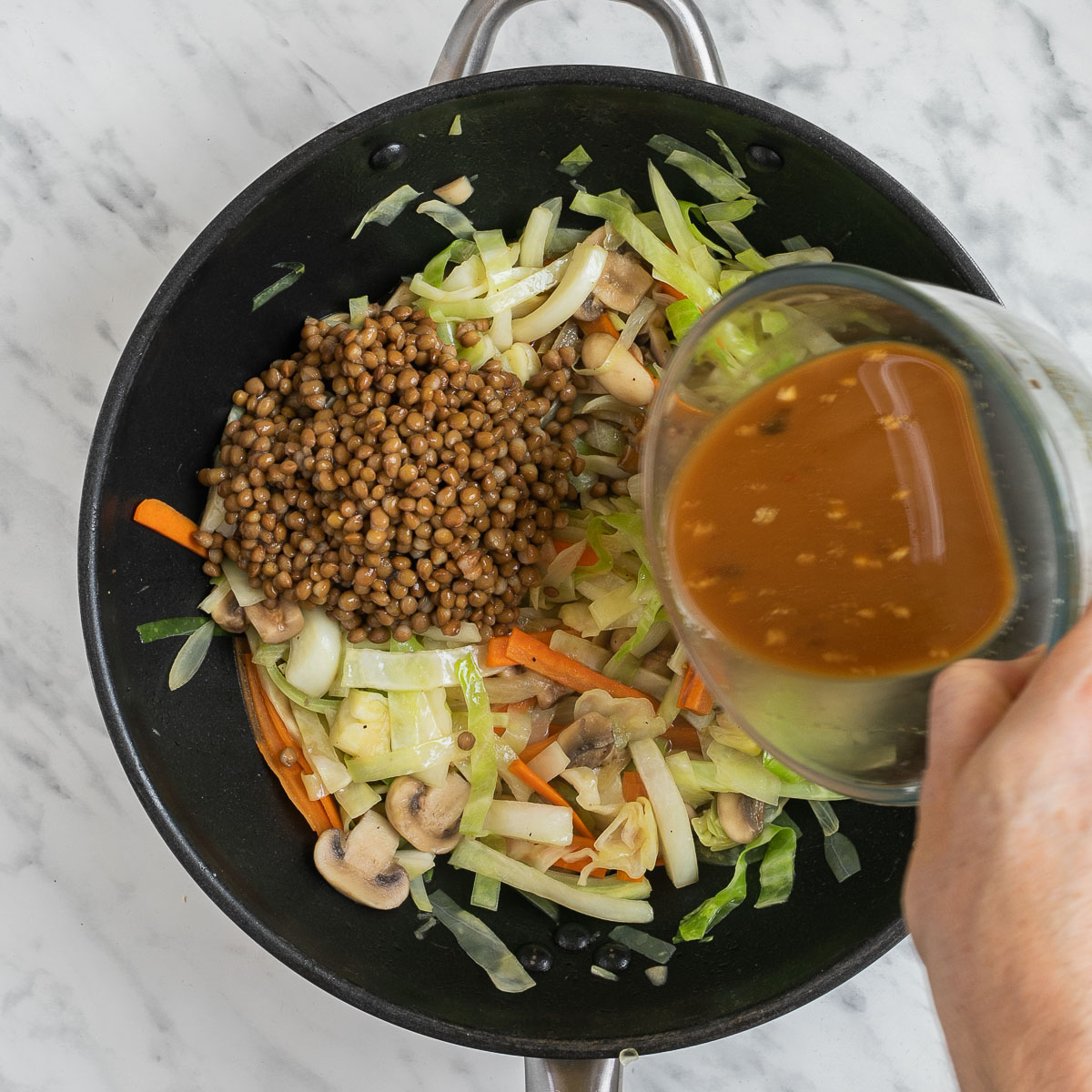 A wok with a mix of sliced mushrooms, shredded cabbage and sliced carrots. A small heap of brown lentils are added. A hand is pouring a sauce on top of them.