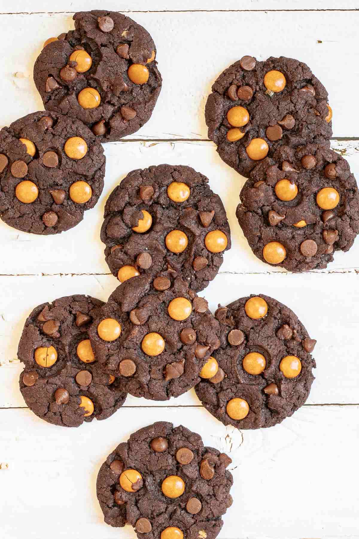 Dark brown cookies with orange m&ms, and chocolate chips on a white wooden surface. A hand is grabbing one.
