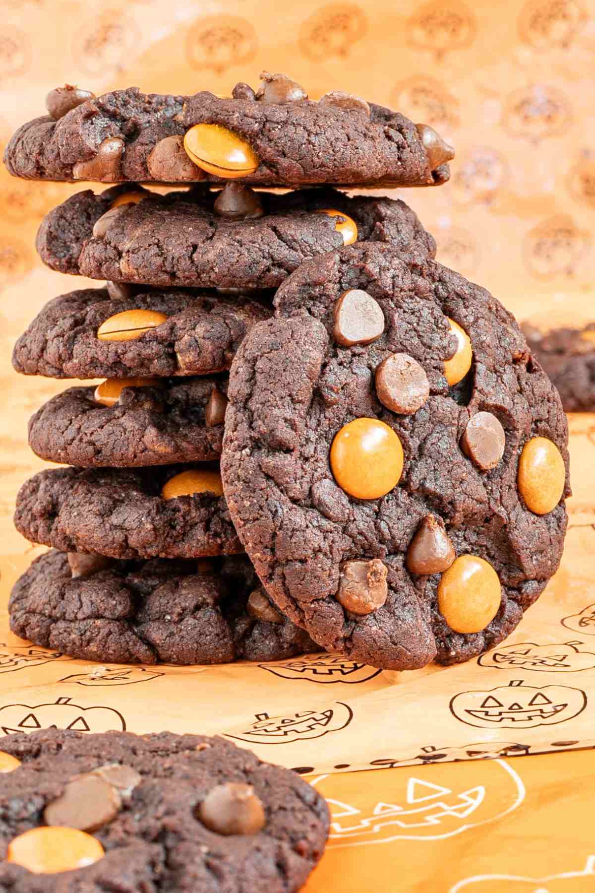 A stack of 6 dark brown cookies with orange m&ms, and chocolate chips on an orange surface with a pumpkin pattern.