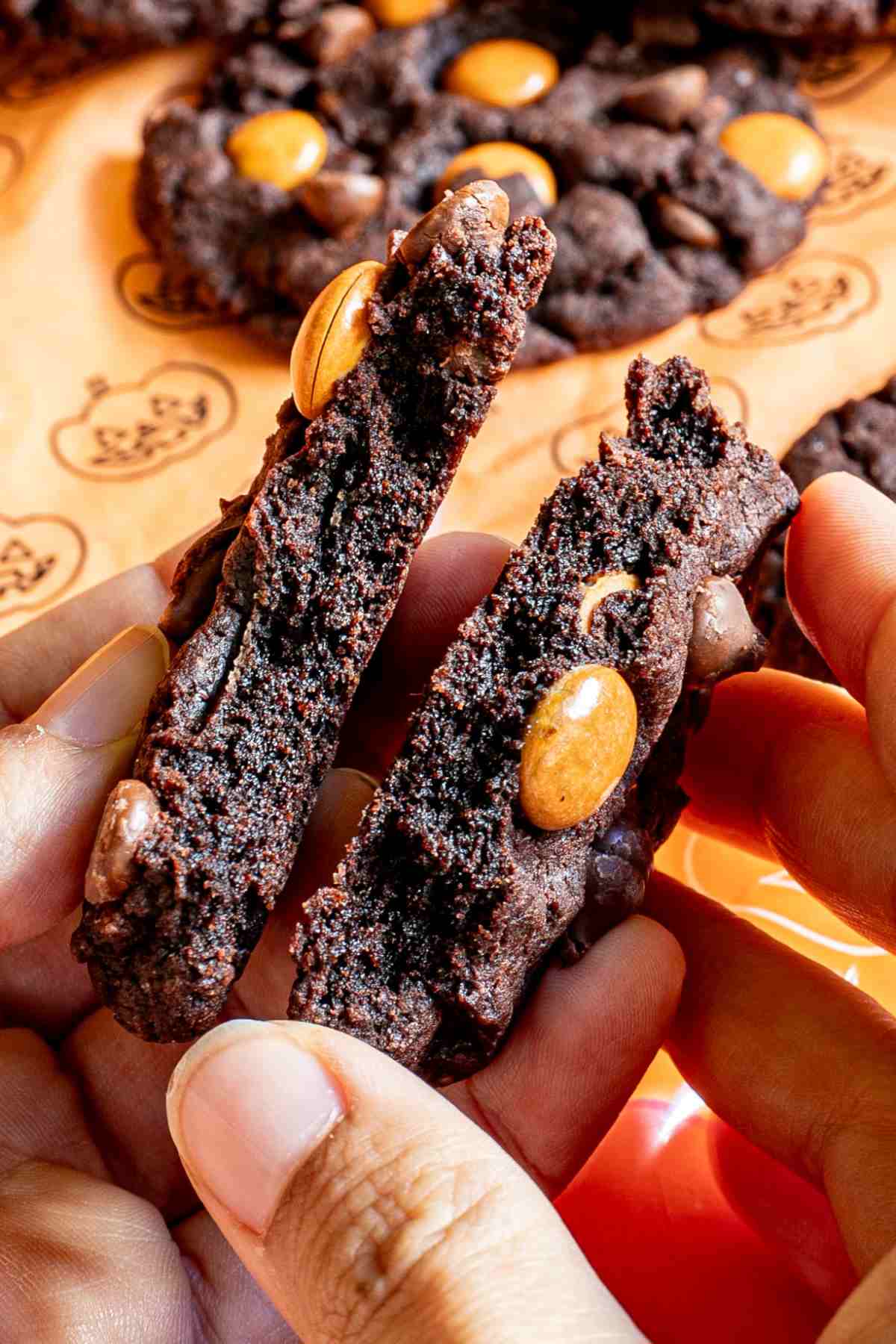 A hand is tearing one dark brown cookie with orange m&ms, and chocolate chips into two halves in front of an orange surface with a pumpkin pattern.