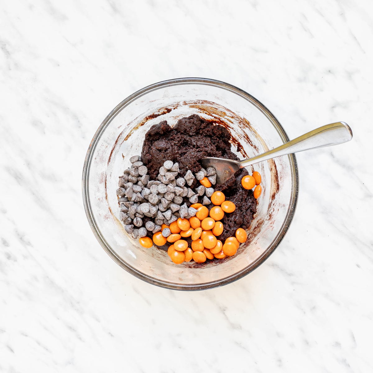 Glass bowl with dark brown cookie dough, orange m&ms, and chocolate chips.