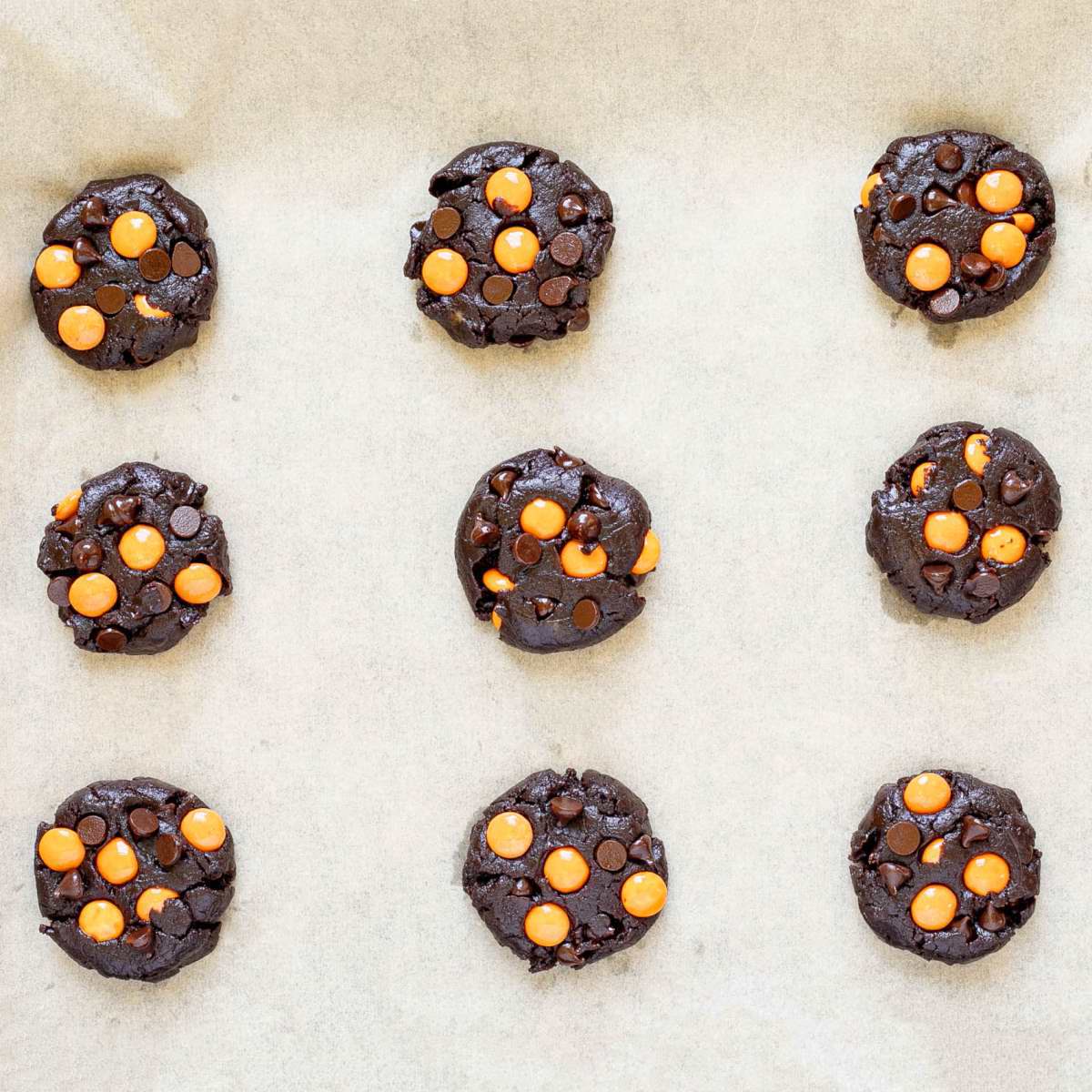Dark brown cookies with orange m&ms, and chocolate chips on parchment paper before baking.
