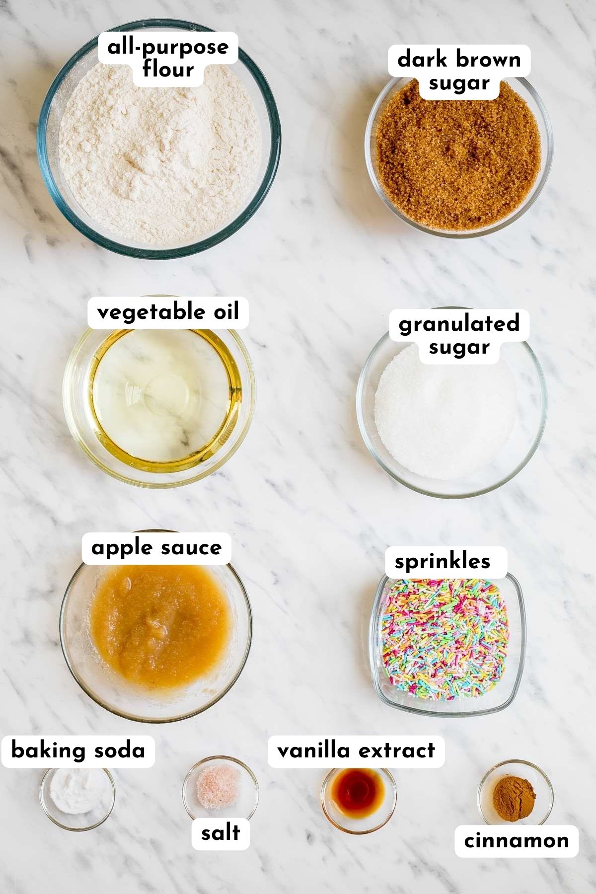 Ingredients of funfetti cookies in small glass bowls like flour, sugar, rainbow sprinkles, apple sauce, oil, salt, cinnamon, vanilla extract, and baking soda.