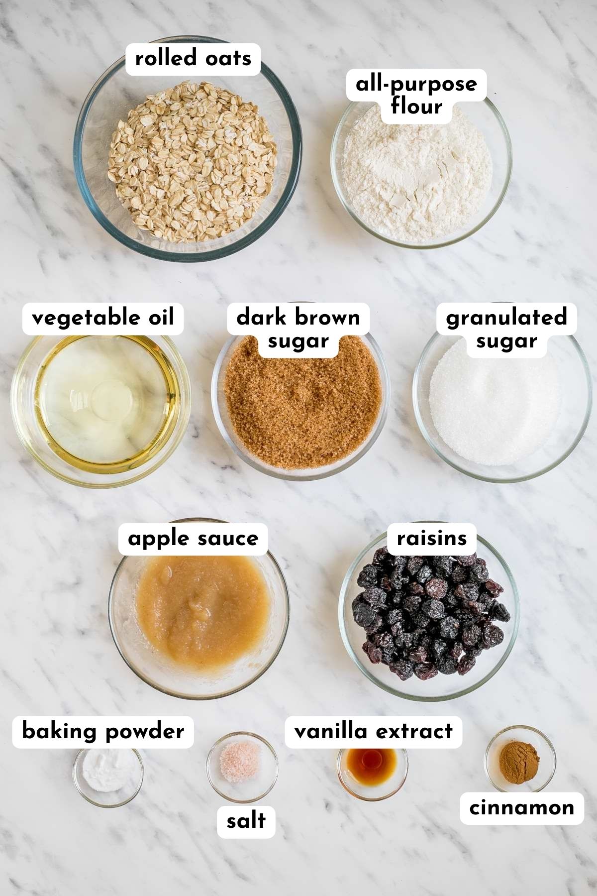 Ingredients of oatmeal raisin cookies in small glass bowls like flour, rolled oats, sugar, raisin, apple sauce, oil, salt, cinnamon, vanilla extract, and baking soda.