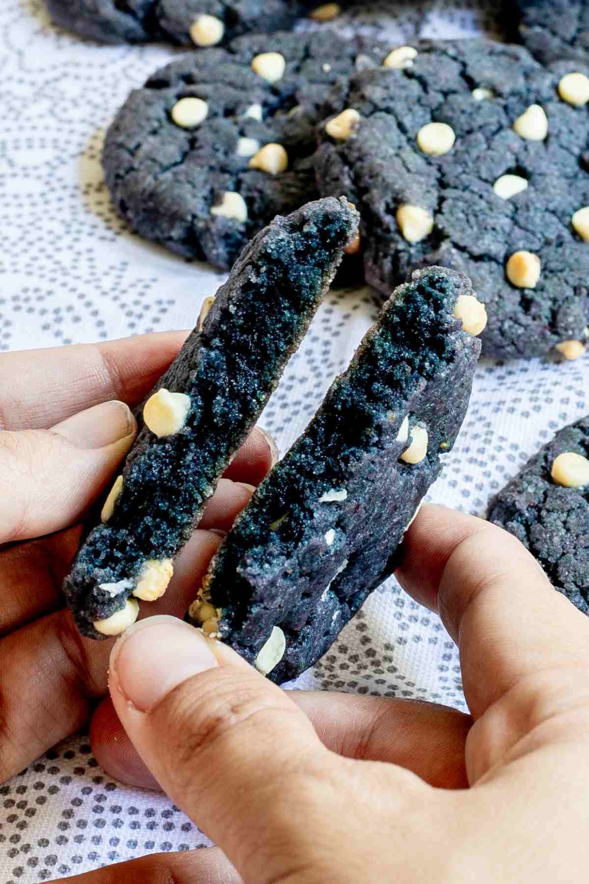 A hand is breaking a blue-purple cookie with white chocolate chips in half to show the inner texture. More cookies are in the background.