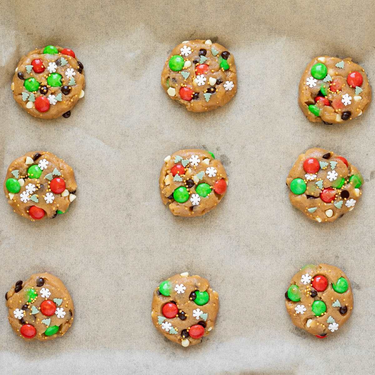 Raw cookie dough with red, green, white and brown candies in them on a parchment paper.