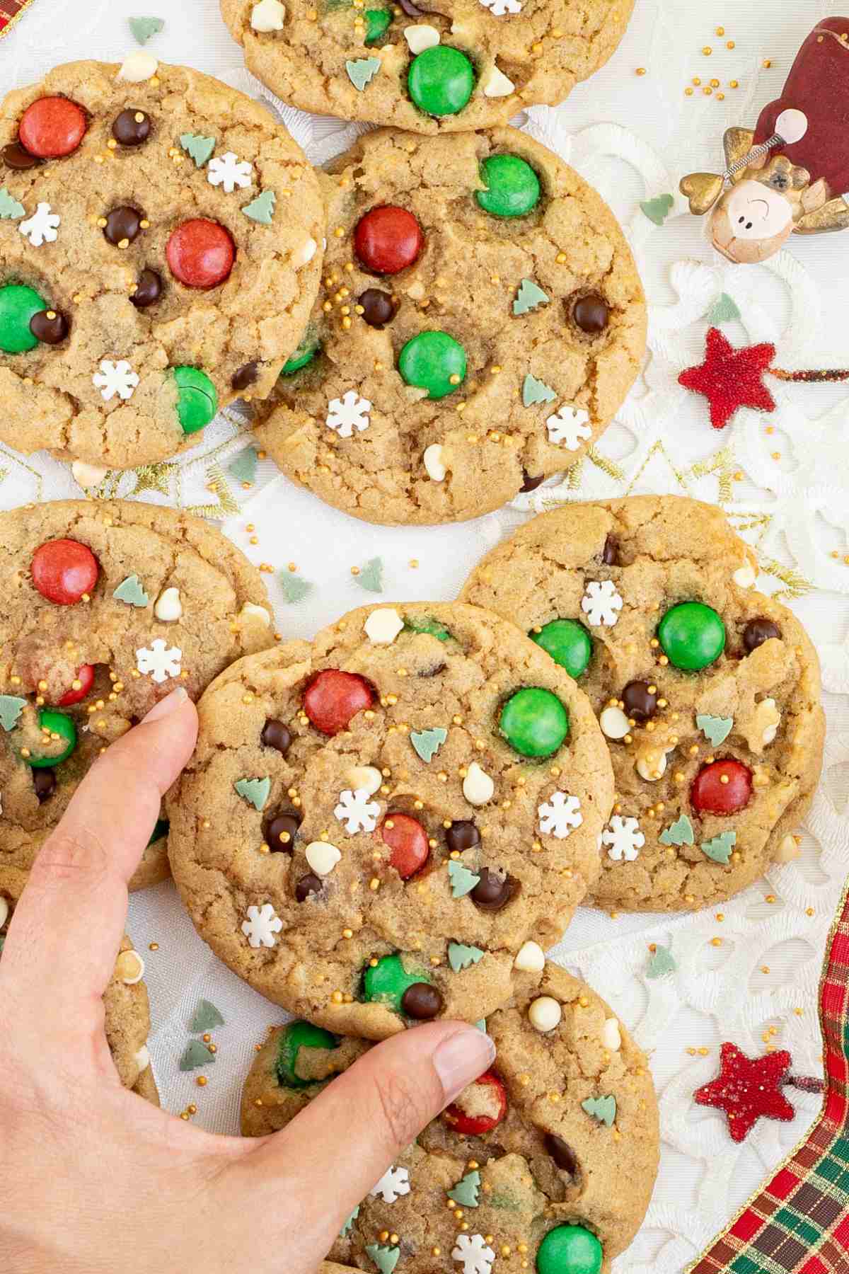 Light brown cookies with red and green m&ms, chocolate chips, tiny white sugar snowflakes, and tiny green pine trees. A hand is grabbing one cookie.