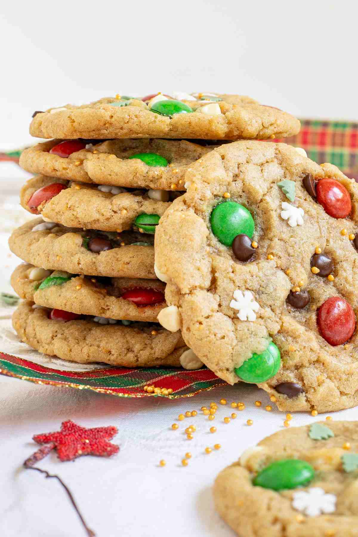 A stack of light brown cookies with red and green m&ms, chocolate chips, tiny white sugar snowflakes, and tiny green pine trees.