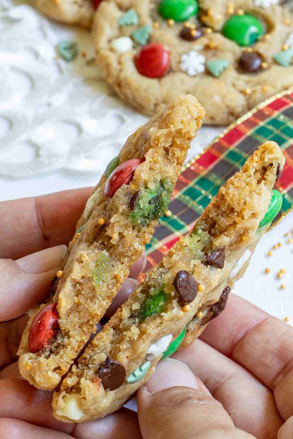 A hand is breaking a light brown cookie with red and green m&ms, chocolate chips, tiny white sugar snowflakes, and tiny green pine trees in half to show the inner texture. More cookies are in the background.