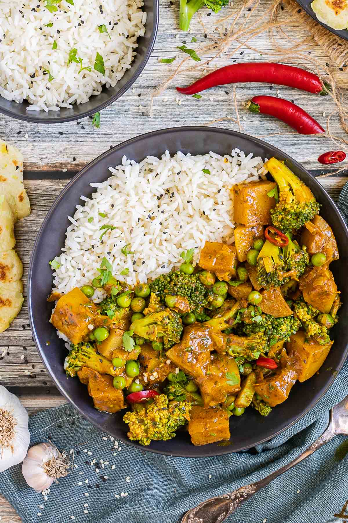 Black plate with rice topped with diced potatoes, broccoli, and green peas glazed in an orange sauce. More rice and flatbreads are placed next to it. 