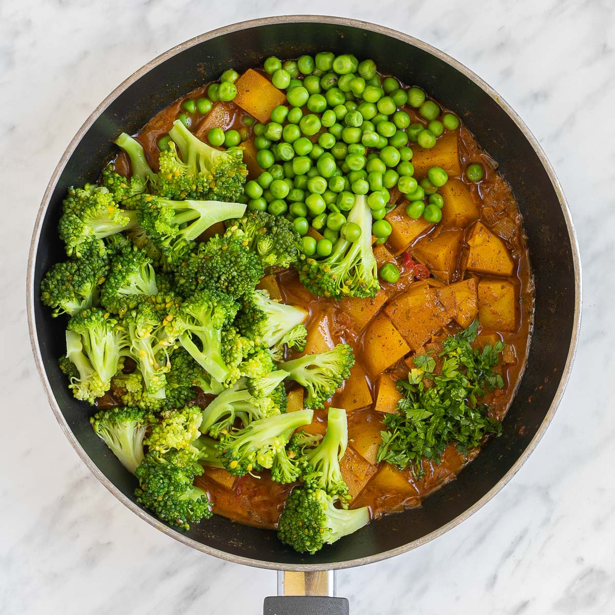 Diced potatoes in an orange sauce in a frying pan topped with broccoli florets and green peas.