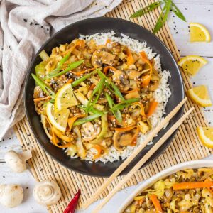 Black plate with white rice topped with stir fired cabbage, mushroom, carrot and lentils. A pair of chopsticks is placed on the side.