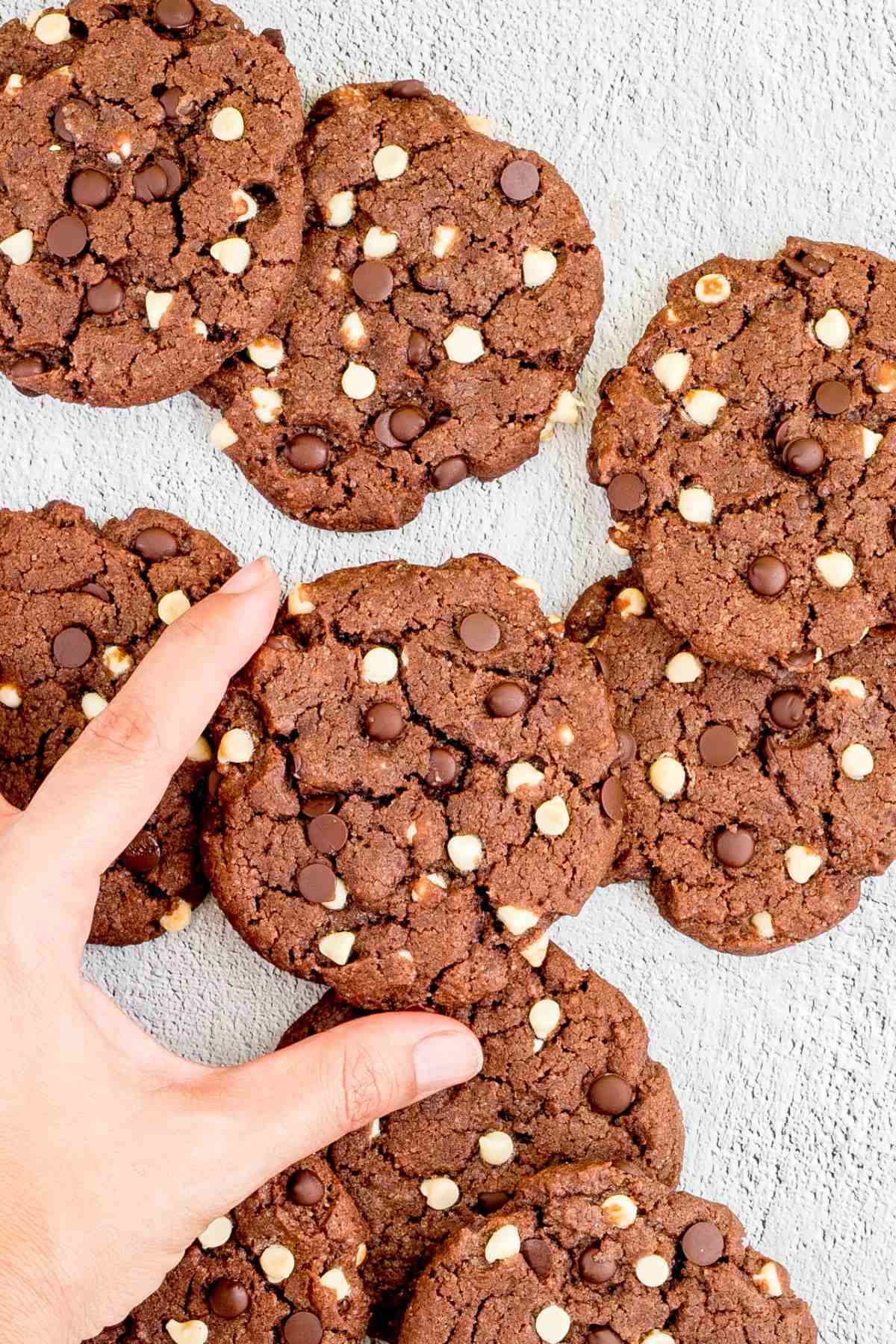 Dark brown cookies with white and dark chocolate chips on a white surface. A hand is taking one from the middle.