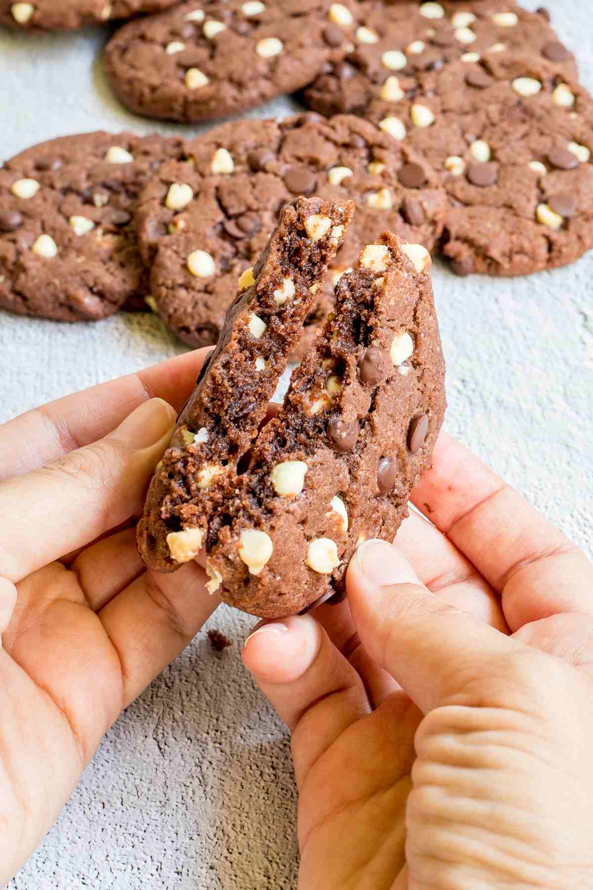 A hand is breaking a dark brown cookie with white and dark chocolate chips in half to show the inner texture. More cookies are behind it.