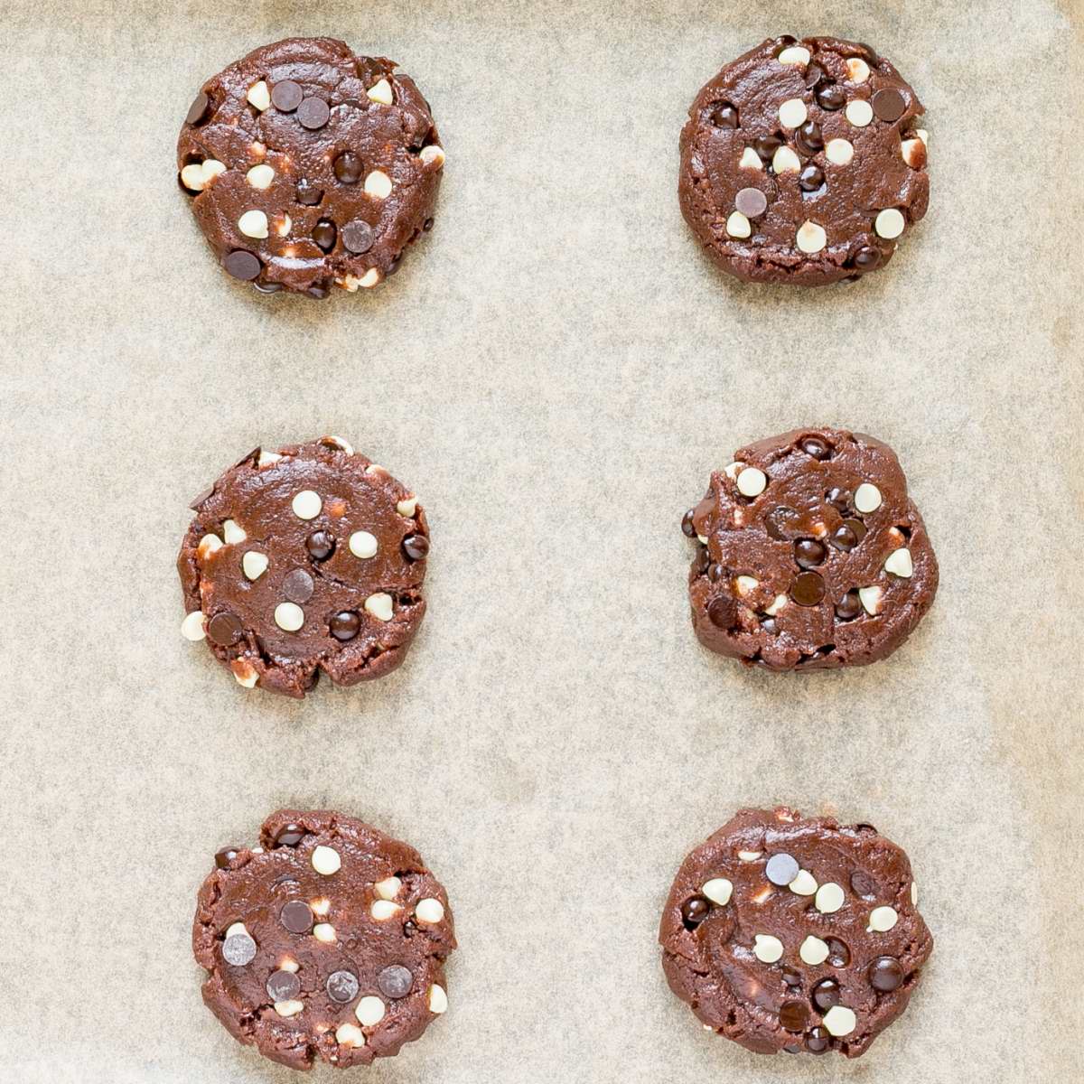 Uncooked dark brown cookies with white and dark chocolate chips on a parchment paper.