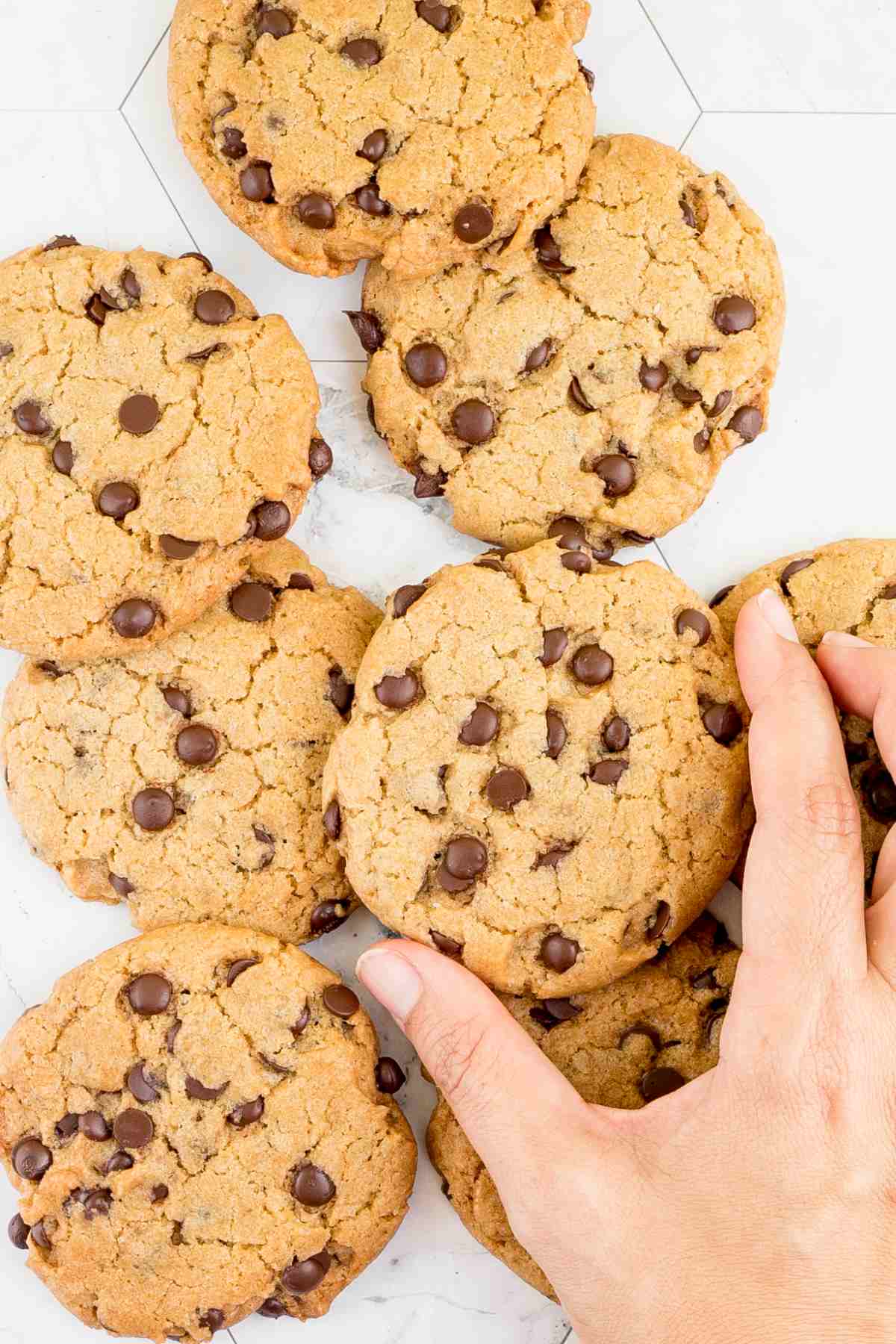 Lots of chocolate chip cookies on a white wooden surface. A hand is about to take one cookie.