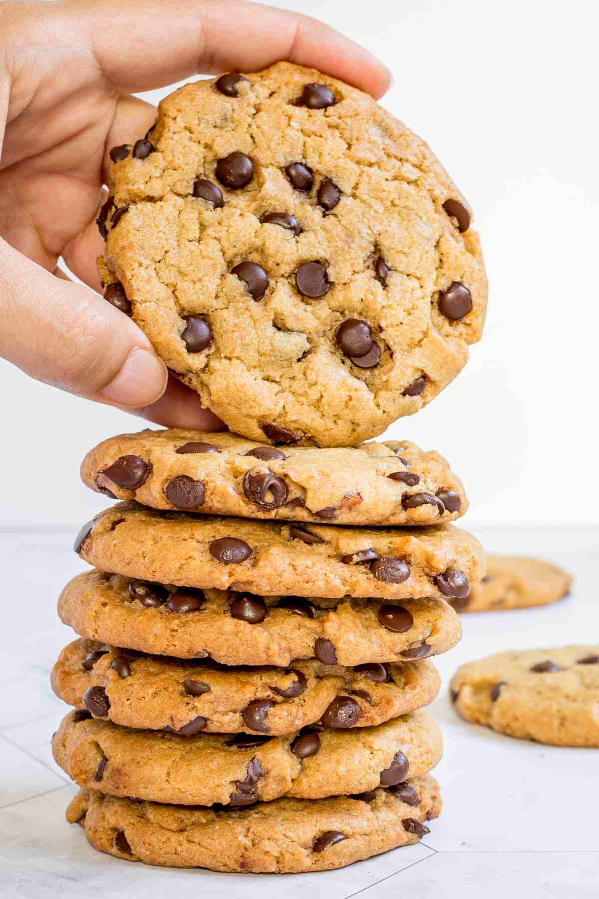 A stack of chocolate chip cookies on a white wooden surface. A hand is taking the top one.